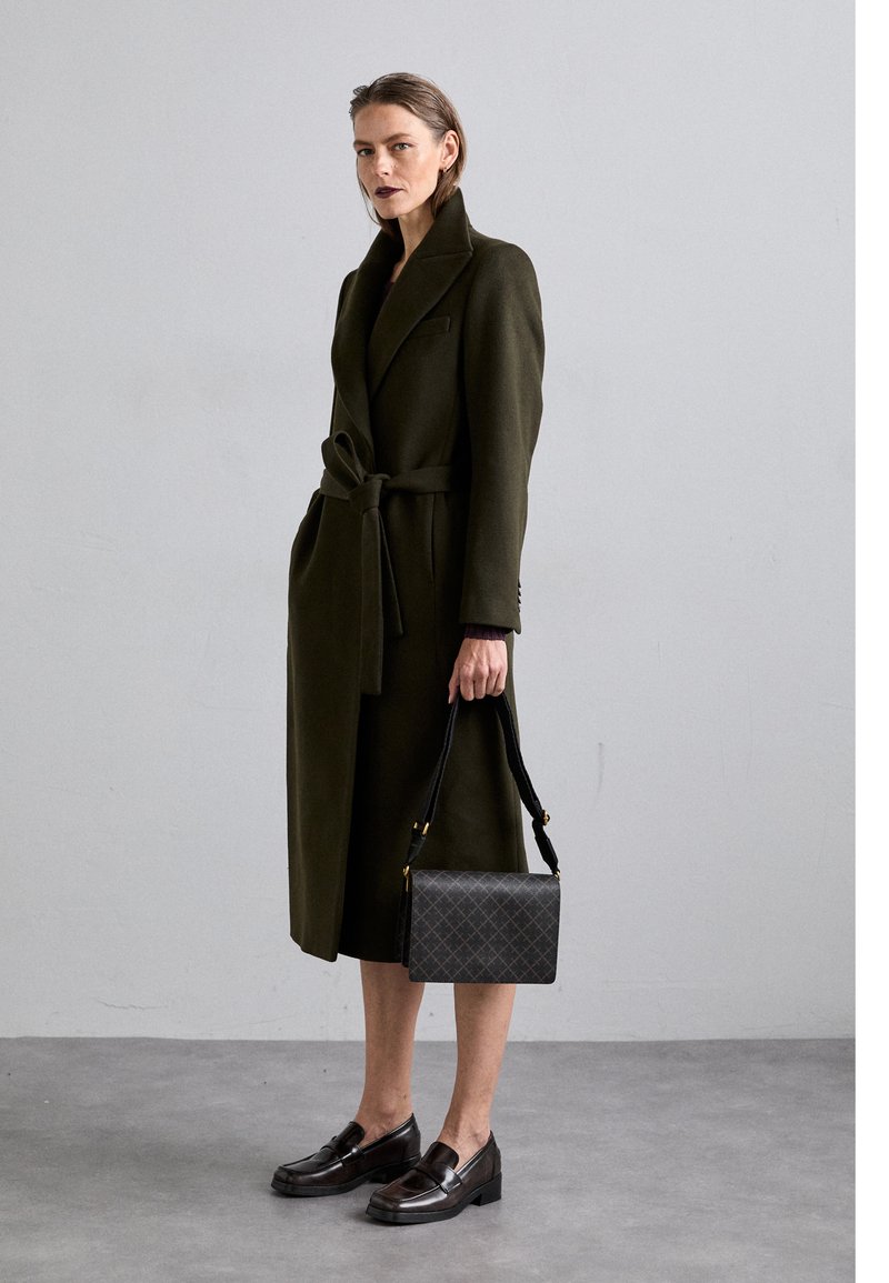 Lång olivgrön kappa med bälte i midjan och serrat krage, kombinerad med en svart mönstrad handväska och mörka loafers, mot en neutral bakgrund.