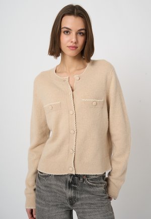 Jonge vrouw met een beige getailleerde vest met knopen en vervaagde zwarte spijkerbroek, staand tegen een effen lichte achtergrond met een neutrale gezichtsuitdrukking.