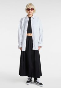 Oversized white button-up shirt with red stitching, black cropped top, black maxi skirt, and black sneakers. Round sunglasses complete the look.