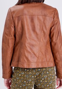 Veste en cuir couleur sable au design ajusté, avec des coutures visibles et une texture lisse. Portée par-dessus un pantalon à motifs verts et jaunes.