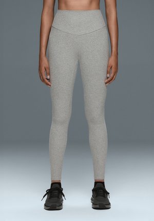 Femme portant des leggings gris clair taille haute et des chaussures de sport noires, debout devant un fond gris uni.