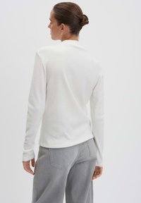 Frau mit einem Dutt, die ein weißes Langarmoberteil und lockere graue Jeans trägt, von hinten vor einem schlichten Hintergrund betrachtet.