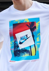 Valkoinen t-paita, jossa on värikäs graafinen kuva palmupuista punaisia ja sinisiä taivasta vasten. T-paidassa on myös vino valkoinen neliö, jossa on Nike-logo ja slogan.