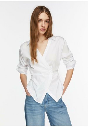 Kobieta o długich brązowych włosach, ubrana w białą bluzkę typu wrap z podwiniętymi rękawami i niebieskie dżinsy, stojąca z rękami w kieszeniach.