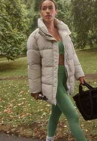 Lys grå pufferjakke med høj krave, parret med en grøn crop top og matchende leggings. Hun holder en vandflaske og en sort tote.