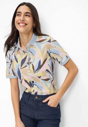 Kvinde smilende, iført en kortærmet polo med blå, beige og gule bladmønstre og mørkeblå jeans, hånd i lommen.