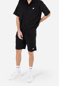 Camicetta nera a maniche corte con bottoni e pantaloni corti abbinati, entrambi in un tessuto texturizzato. Logo bianco sulla camicia e sui pantaloni. Sneakers bianche e calzini.