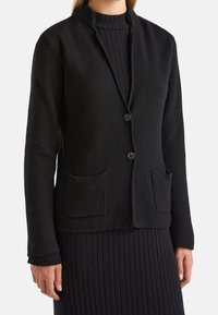 Černý pletený cardigan s texturovanou tkaninou, zapínání na jeden knoflík a dvěma předními kapsami. Obsahuje límec mandarínského stylu a žebrované manžety.