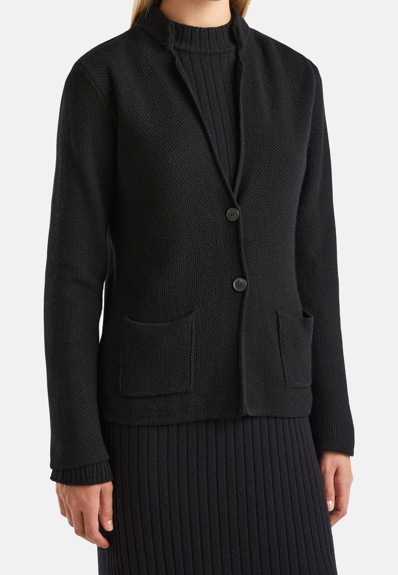 Černý pletený cardigan s texturovanou tkaninou, zapínání na jeden knoflík a dvěma předními kapsami. Obsahuje límec mandarínského stylu a žebrované manžety.