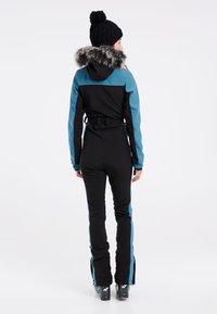 Zwarte en teal skibroek met een nepbont afgewerkt capuchon, strakke pasvorm en ritssluitingen bij de enkels; gedragen met een zwarte gebreide beanie.