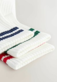 Trois chaussettes blanches en maille côtelée pliées, avec des rayures rouges, vertes et bleu marine aux bords, empilées sur une surface claire.