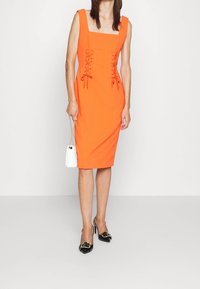 Oranžové upnuté šaty se čtvercovým výstřihem a šněrovacími detaily v pase, doplněné černými podpatky a bílou kabelkou.