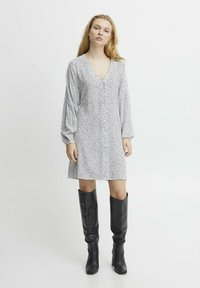 Weißes Kleid mit schwarzen Sprenkeln, V-Ausschnitt und langen Puffärmeln, kombiniert mit kniehohen schwarzen Lederstiefeln, die einen kontrastierenden Look kreieren.