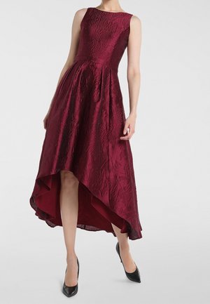 Ärmelloses, strukturiertes bordeauxrotes Kleid mit High-Low-Saum, kombiniert mit schwarzen spitzen High Heels auf neutralem Hintergrund.