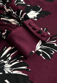 Burgundy kukkaistuinen mekko lähikuvassa, jossa on mustavalkoisia kukkakuosisia, pehmeä rakenne ja nappidetaili hihassa.