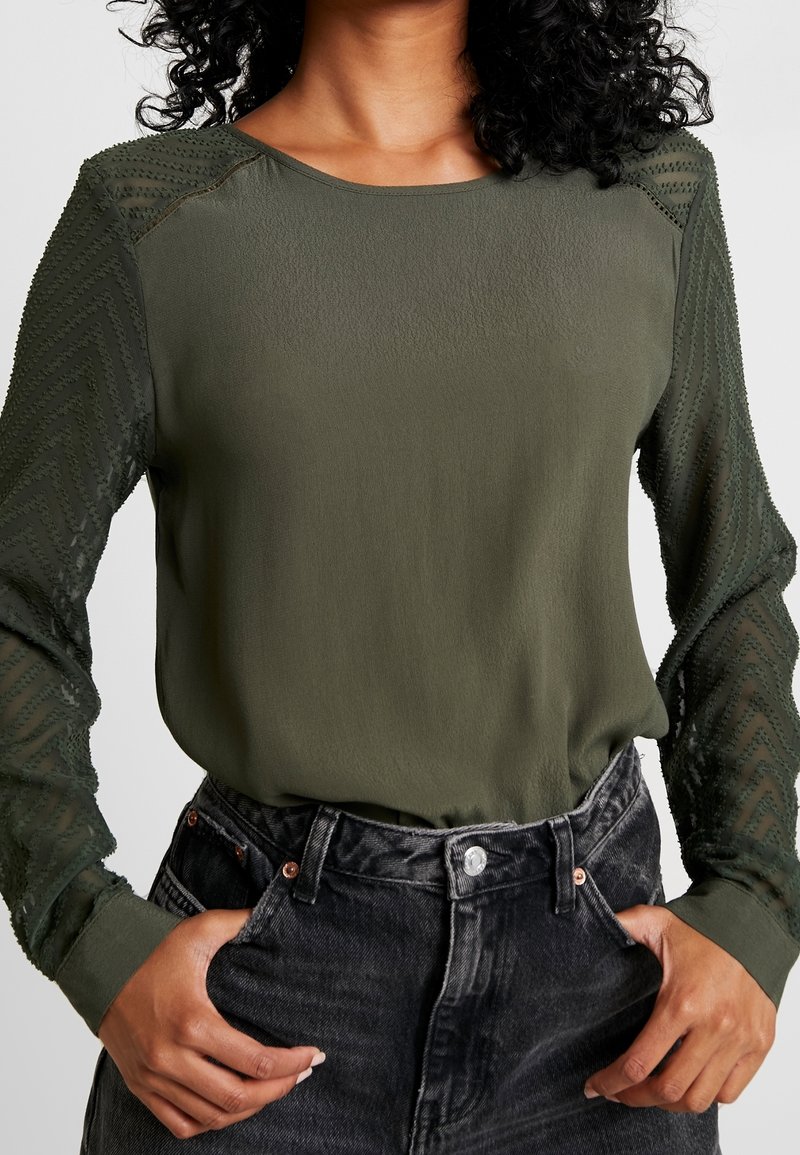 Donna che indossa una blusa verde oliva con maniche trasparenti texturizzate, infilata in jeans neri sbiaditi a vita alta, con le mani nelle tasche.