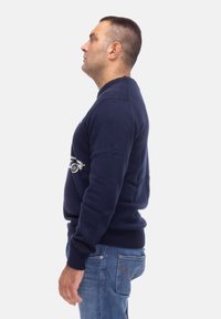 Maglione blu navy con scollatura rotonda, polsini a costine e design del logo. Indossato con jeans azzurri. Maniche corte; trama uniforme.