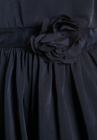 Navy blue dress featuring a textured fabric with pleats and a floral fabric accent at the waist, detailed with a smooth satin ribbon.