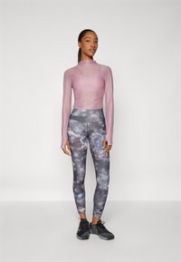 Eine stehende Frau trägt ein durchsichtiges, langärmeliges mauvefarbendes Oberteil über einem passenden Sport-BH, mehrfarbige gemusterte Leggings und schwarze Sneaker.