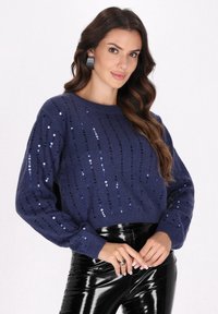 Pulover tricotat bleumarin, cu paiete în dungi verticale, având un guler rotund și mâneci lungi, asortat cu pantaloni negri lucioși.