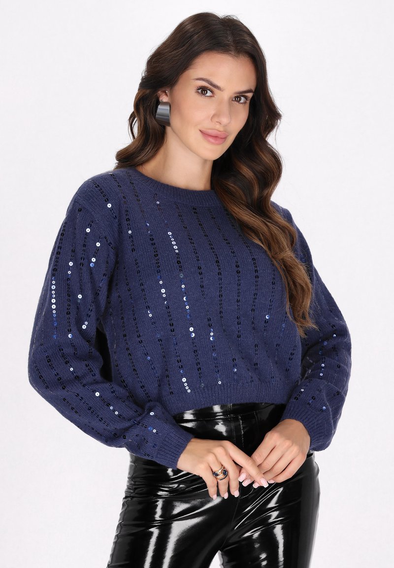 Pulover tricotat bleumarin, cu paiete în dungi verticale, având un guler rotund și mâneci lungi, asortat cu pantaloni negri lucioși.