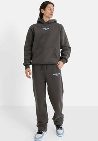 Donkergrijze hoodie en bijpassende joggingbroekset, met een buidelzak aan de voorkant, minimalistische blauwe logotekst en geribbelde manchetten, gecombineerd met lichte sneakers.