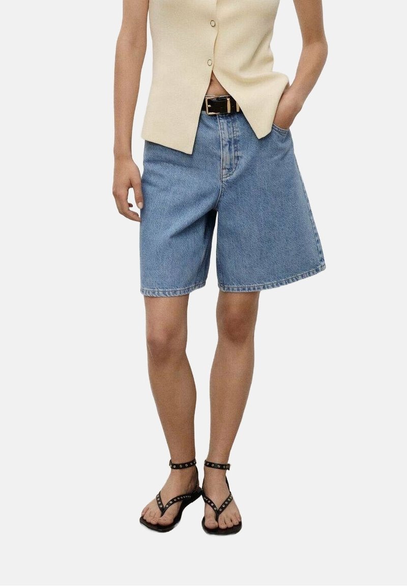 Personne portant un short en jean bleu clair avec une ceinture noire, un haut beige sans manches et des sandales noires cloutées.