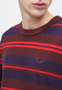 Πλεκτό πουλόβερ σε υφή βάφλας με οριζόντιες ρίγες σε μπορντό, ναυτικό μπλε και κόκκινο. Διαθέτει ένα μικρό, κυκλικό λογότυπο στο στήθος.