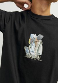 Camiseta de algodón negra con un estampado en acuarela de botellas de vidrio, con el texto "ATELIER Originals™ STUDIO ©1990" en color crema.