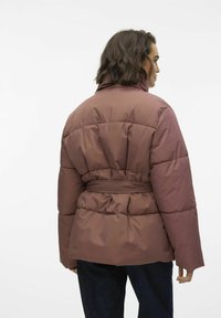 En pufferjakke i en dempet brunfarge med belte i midjen, quiltet tekstur, bred krage og lange ermer. Designet er oversized og uformelt.