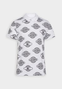 Vit pikétröja med ett upprepande mönster av svart logotyp med globgrafik och texten "LACOSTE". Inkluderar klassisk krage och korta ärmar.