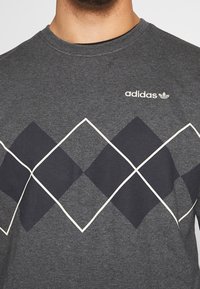 Grå bomull T-shirt med ett stort mörkt diamanternötlägg med vita konturer. Inkluderar en liten vit "adidas"-logotyp vid bröstet.