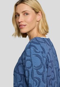 Blaues strukturiertes Sweatshirt mit einem erhöhten, kreisförmigen Muster, das einen runden Halsausschnitt und einen dezenten Glanz aufweist. Kabelstrick-Akzente auf der Schulter.