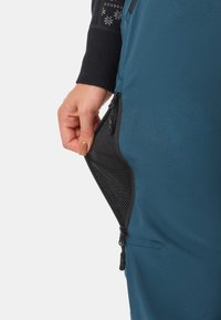 Käsi öppnar en sidficka med svart mesh-foder på blå byxor, visar dragkedjedetaljer och en del av en svart långärmad topp.