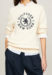 Cream sweater with ribbed texture, featuring a large dark logo and laurel design. Worn over a blue collared shirt, with rolled cuffs.