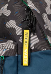 Lo zaino presenta un tessuto grigio con un motivo e dettagli blu, un fondo turchese e un'etichetta gialla "RESISTI ALLA SPRECO". Include zip resistenti.