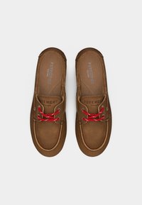 Καφέ παπούτσια στυλ μοκασίνου με κόκκινα κορδόνια, τοποθετημένα επίπεδα, με ραμμένες ραφές και ορατές εσωτερικές σόλες με λογότυπο.