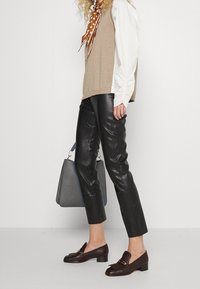 Gilet en tricot brun sur chemise blanche, pantalon en cuir noir, mocassins bruns avec des accessoires en or, tenant un sac à main gris. Textures lisses et design ajusté.