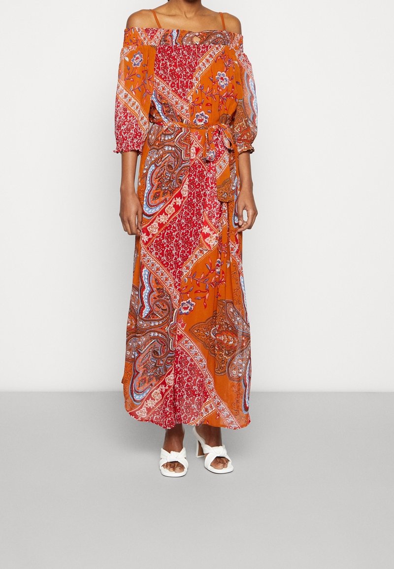 Femme portant une longue robe épaules dénudées avec des motifs orange, rouge et cachemire, et des sandales blanches à talons ouverts.