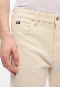 Gros plan sur la main gauche d'une personne reposant près de la poche d'un jean couleur crème avec une étiquette noire "BOSS".