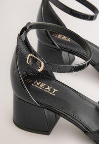 Sandales à talon carré en cuir verni noir avec bride à la cheville. Présentent un marquage doré sur la semelle intérieure et un côté texturé pour une meilleure adhérence.