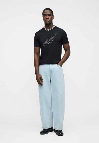 Άνδρας στέκεται φορώντας μαύρο γραφιστικό μπλουζάκι με ριγέ γιακά, ανοιχτό μπλε τζιν με φαρδιά πόδια, μαύρα παπούτσια, χέρι στην τσέπη, ουδέτερο φόντο.