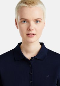 Poloko námořnické modré barvy vyrobené z texturované látky, s klasickým límcem, zapínáním na knoflíky a malým vyšitým logem na levé hrudi.