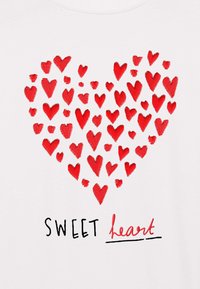 Balta marškinėliai su dideliu raudonu širdies dizainu, kuriame yra įvairių dydžių širdelių ir tekstu "SWEET heart" juoda ir raudona spalvomis žemiau.