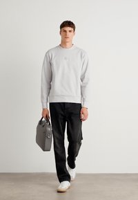 Felpa grigia chiara con collo rotondo, jeans neri e sneakers bianche, mentre tiene una borsa a tracolla grigia. Design semplice, accenti minimi.