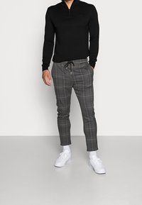 Svart zip-up tröja med långa ärmar, parat med grå rutiga tapered byxor med dragsko i midjan och vita sneakers.