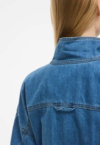 Denim jakna v srednje modri barvi, z oblikovanim ovratnikom in šivom na hrbtni strani. Tekstura tkanine se zdi trdna in mehka, z vidnimi šivi.