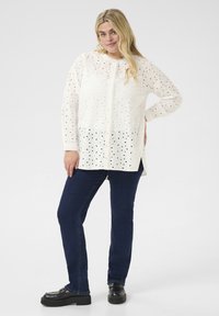 Blusa blanca de ojal con cierre de botones, con recortes florales y mangas largas, acompañada de vaqueros de denim azul oscuro y zapatos de plataforma negros.