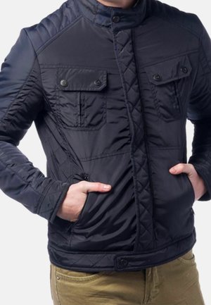 Mann trägt eine dunkelblaue gesteppte Jacke mit aufgesetzten Brusttaschen und Knöpfen sowie die Hände in den Seitentaschen, kombiniert mit hellbraunen Hosen.