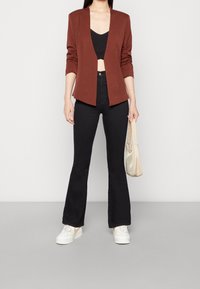 Hnědý blazer s otevřeným předním dílem, černý crop top, černé rozšířené džíny a bílé tenisky. Drží světlou tašku. Jednoduchý design a textury.
